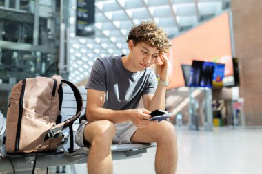 Young man with backpack sitting at airport looking worried at passport and smartphone. Represents stress, flight delay, and challenges of travel lifestyle