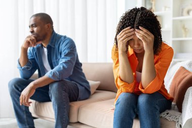 Üzgün Afrikalı Amerikalı çift evdeki kanepede yan yana oturuyor, kadın stres içinde başlarını tutuyor. İlişki sorunları, anlaşmazlık, gerginlik, yaşam tarzı ve aile kavramı.