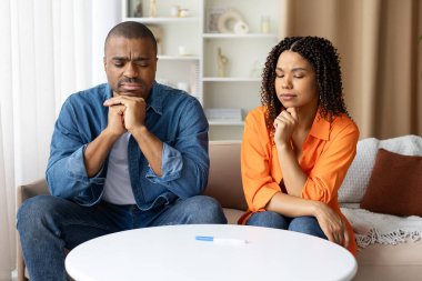 Endişeli Afrikalı Amerikalı çift masada oturmuş hamilelik testine bakıyor, hem ciddi hem de duygusal. Aile planlaması, stres, belirsizlik ve yaşam tarzı kavramı