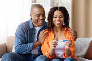 Gülümseyen Afrikalı Amerikalı çift evde pozitif hamilelik testine bakıyor. Aile planlaması, neşe, aşk, yaşam tarzı, çocuk ve mutluluk beklentisi