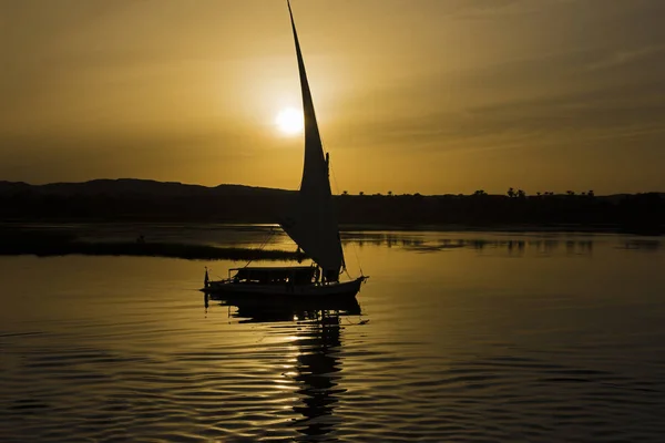 アスワンで夕方には夕暮れのオレンジ色の夕日の反射とナイル川の伝統的なエジプト フェルッカ航行ボートのシルエット — ストック写真