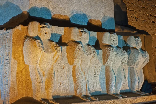 Statues on base of large obelisk at entrance pylon to ancient egyptian Luxor Temple lit up during night