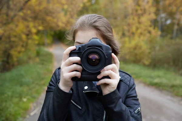 一个金发碧眼的年轻女孩在皮革夹克看进入取景器的专业相机 准备拍摄一个美丽的分期广告 或报告文学框架 对模糊的沥青路面 绿色的草与小 Num — 图库照片