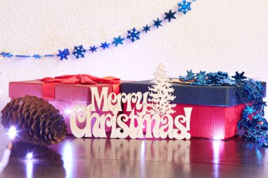 Neşeli Noel-poster veya kartpostal tasarım. Hangi ahşap bir yazıt, takı, hediyeler 2 kutu ve bir çam kozalağı, Festival çelenk gösterir. Bütün bu beyaz bir duvar karşı ahşap bir masaya yatıyor