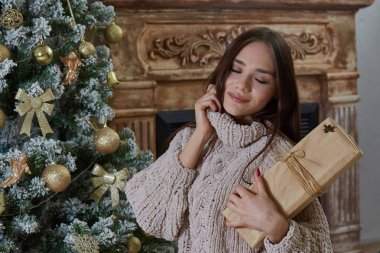 Kız tutar bir Noel hediyesi ve hayal büyülü hediyeler, gözleri kapalı ve Yaka Kazak, şömine ve tatil dekorasyon ile Noel ağacı arka plan üzerinde tutarak, o bir bej, Kraft kağıt sarılmış hediye
