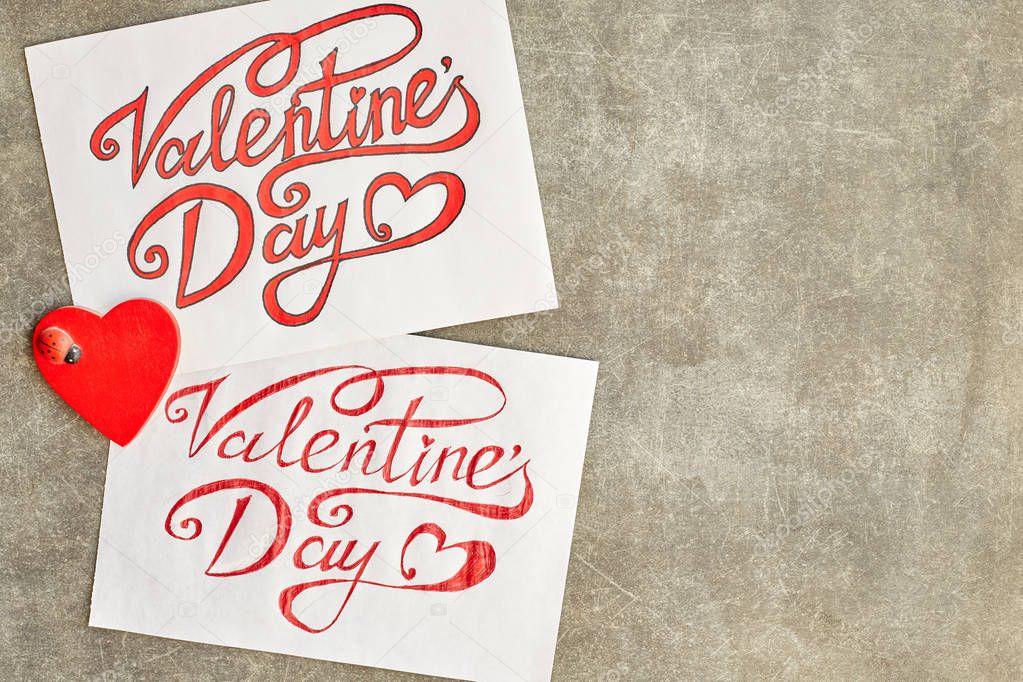 Paper sheets with a painted inscription Valentine's Day beautiful font, red. On one painted in ink and gently on the other red gel pen, less carefully, between them a wooden red heart with a ladybug. They lie on the old scratched up gray background.