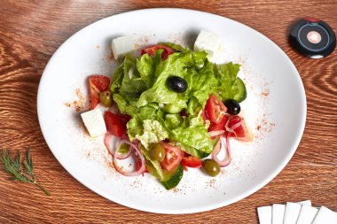 salatalık, domates, zeytin, soğan, marul, mozzarella salatası yatıyor beyaz peçeteler, dereotu ve düğme delikanlı ile ahşap bir masa üzerinde duran kırmızı biber ile süslenmiş beyaz bir plaka üzerinde