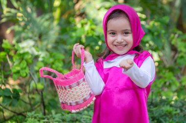 Güzel bir kız, beyaz, karizmatik, kahverengi saçlı, siyah gözlü, üç yaşında, pembe giyinmiş, masha tarzı, çok etkileyici, bahçede yürüyor, oyuncak ayı ve çantayla oynuyor. Yeşil çimenler ve ağaçlar