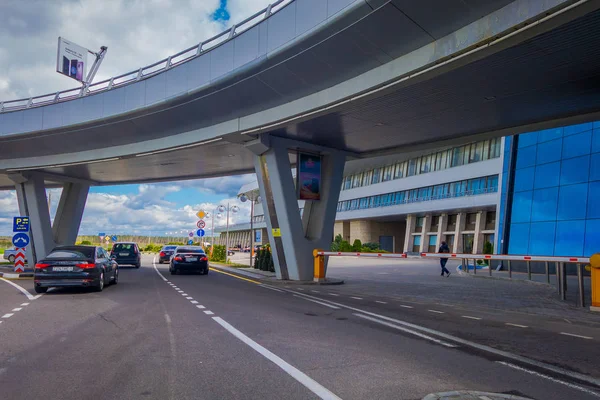 MINSK, BELARUS - 01 DE MAYO DE 2018: Vista de personas caminando y coches estacionados en la entrada del edificio del aeropuerto de Minsk en un día nublado con una estructura de puente apedreado iat la entrada del aeropuerto en Bielorrusia —  Fotos de Stock