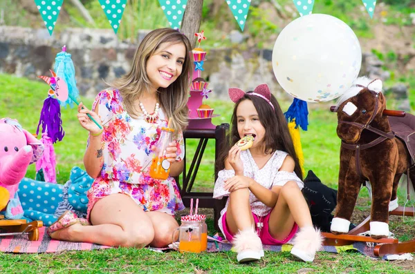 Wanita tersenyum tak dikenal dengan botol jus di tangan bersama putri kecilnya yang sedang makan kue, mengelilingi pesta di taman pada hari musim panas yang cerah — Stok Foto