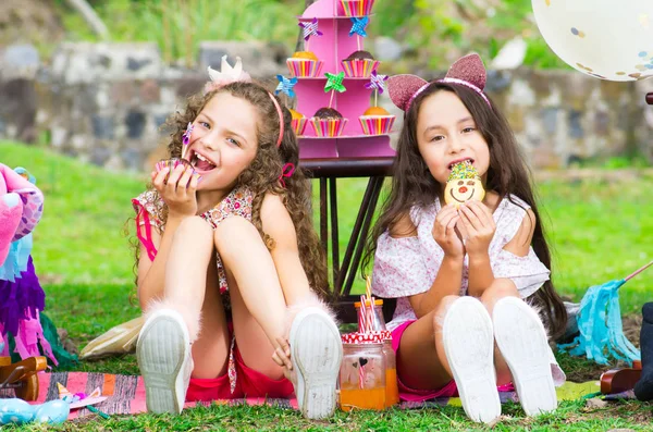 Dos monas hermanas en el jardín, rizada comiéndose una magdalena y caucásica comiendo una galleta al aire libre, en un soleado día de verano vistiendo ropa de colores pastel —  Fotos de Stock