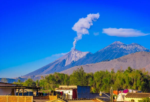 Bellissimo paesaggio di montagna enorme in procinto di aruption con una colonna di cenere, vista dai tetti dell'edificio nella città di Antigua in splendida giornata di sole e cielo blu — Foto Stock