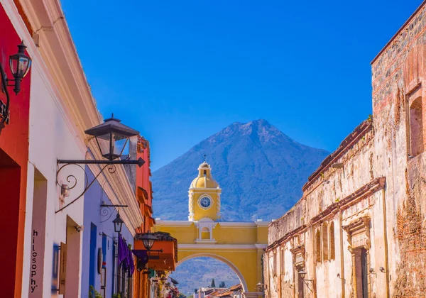 Ciudad de Guatemala, Guatemala, 25 aprile 2018: Veduta esterna della vecchia strada con edifici clasici nella città di Antigua con arco giallo colorato con il vulcano Agua attivo sullo sfondo — Foto Stock