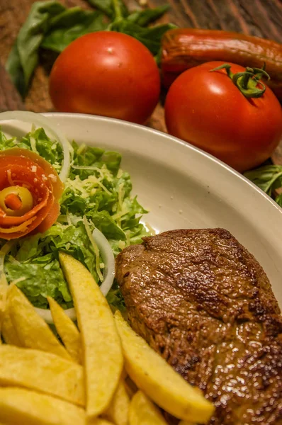 Primer plano del detallado filete a la parrilla bien hecho de Nueva York con papas fritas, tomates, salchichas. servido en placa blanca sobre tabla de cortar sobre fondo de mesa de madera —  Fotos de Stock