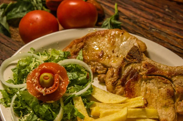 Вкусная отбивная с картошкой фри, помидорами и салатом подается в белой тарелке на деревянном фоне стола — стоковое фото