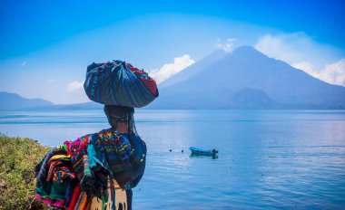 Panajachel, Guatemala-Nisan, 25, 2018: tipik giyim eşyası ve içinde göl kıyısındaki küçük teknelerle Atitlan göl ve arka planda yanardağ ile yürüme unidentifed yerli kadın, açık görünüm