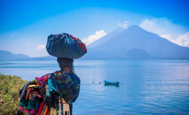 Panajachel, Guatemala -April, 25, 2018: Outdoor view of unidentifed indigenous woman, wearing typical clothes and walking in lakeshore with small boats in Atitlan Lake and volcano in Background in clipart