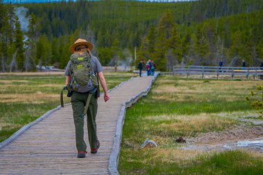 Yellowstone, Montana, ABD 24 Mayıs 2018: Açık yeşil bir sırt çantasıyla, üniforma giyen kadın park korucusu eski sadık üst şofben havzasında ahşap yol boyunca yürüyüş görünümünü