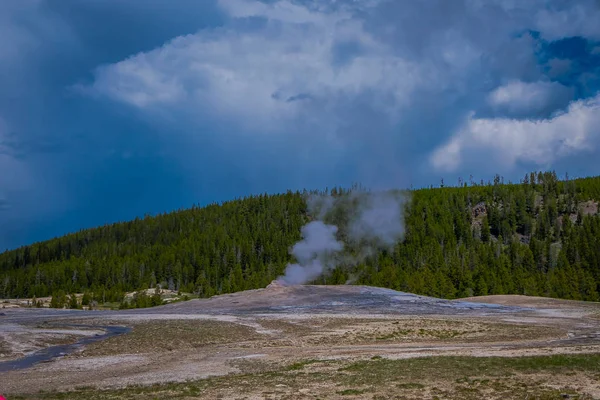 Hermosa vista de la antigua y fiel cuenca del géiser ubicada en el Parque Nacional de Yellowstone, rodeada de vapor con una montaña verde detrás en un hermoso día soleado — Foto de Stock