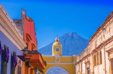 Ciudad de Guatemala, Guatemala, April, 25, 2018: Outdoor view of old street with clasical buildings in the city of Antigua with colorful yellow arch with the active Agua volcano in the background clipart