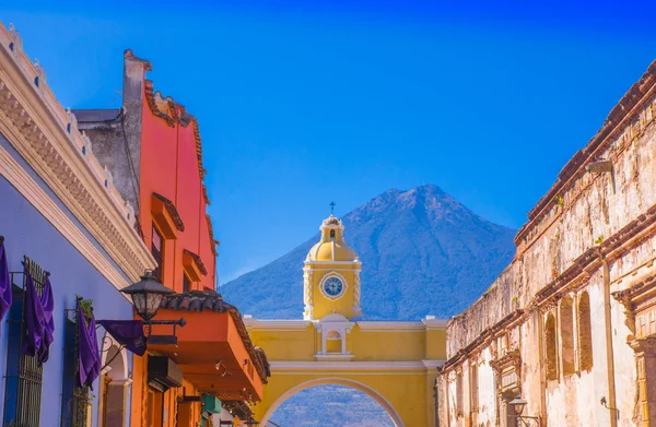 Ciudad de Guatemala, Guatemala, April, 2018: Outdoor view of old street with clasical buildings in the city of Antigua with colorful yellow arch with the active Agua volcano in the background — стоковое фото