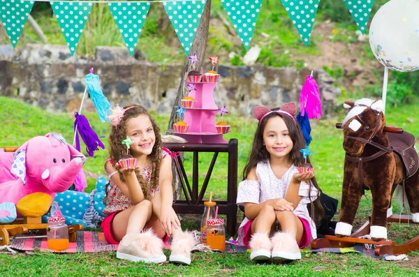 Две красивые белые маленькие сестры держат в руках вкусный кекс на открытом воздухе, в парке в солнечный летний день носить пастельные цвета одежды с партийной начинкой — стоковое фото