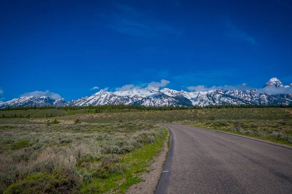 Vista ao ar livre da rodovia apedrejada que leva ao Grand Teton National Park, Wyoming, em um lindo dia ensolarado com montanhas cobertas de neve na horizontal — Fotografia de Stock