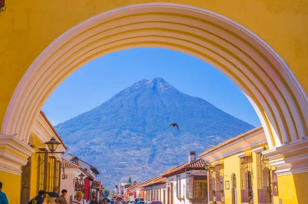 Ciudad de Guatemala, Guatemala, 25 апреля 2018 года: Неизвестные ходят по улицам города Антигуа и смотрят на активный вулкан Агуа на заднем плане через цветную желтую арку — стоковое фото