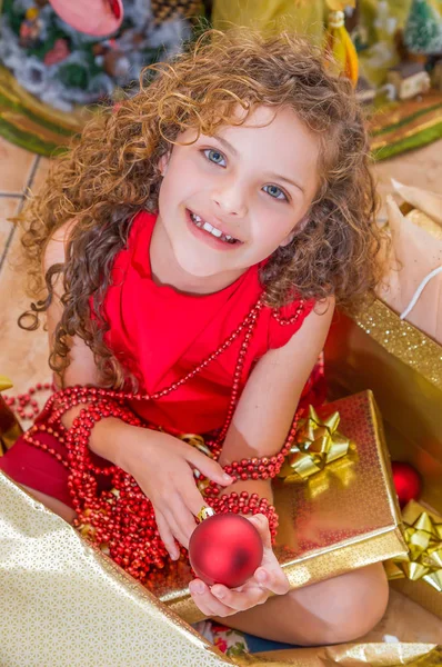 Primo piano di una ragazza felice che indossa un vestito rosso e tiene in mano un ballo di Natale e una decorazione, con un albero di Natale dietro, concetto di Natale — Foto Stock