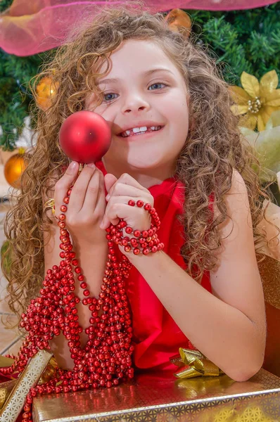 Gros plan de fille heureuse portant une robe rouge et tenant une boule de Noël et la décoration dans ses mains, avec un arbre de Noël derrière, concept de Noël — Photo