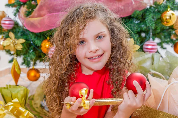 Primo piano della ragazza sorridente che indossa una camicetta rossa e tiene in mano una palla di Natale, una decorazione e un regalo, con un albero di Natale dietro, concetto di Natale — Foto Stock