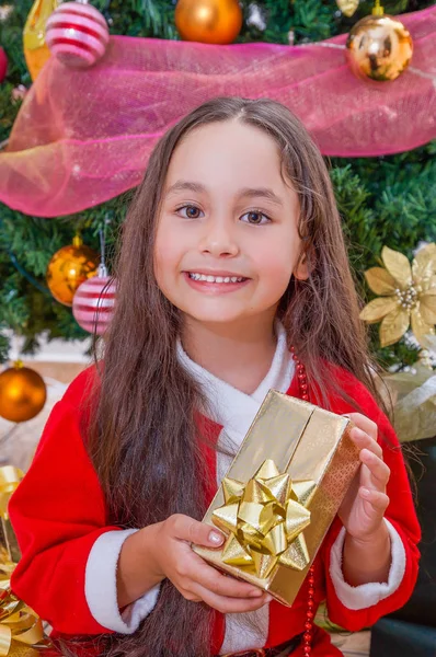 Close up van lachende meisje draagt een rode santa kostuum en bedrijf van een gift in haar handen, met een kerstboom achter, Kerstmis concept — Stockfoto