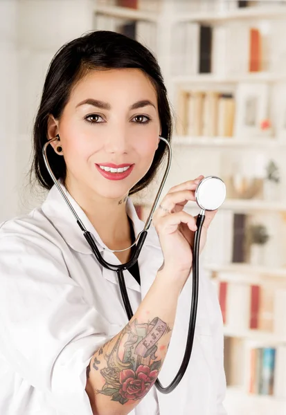 Beautiful smiling tattooed young doctor holding the chest part of the stethoscope in her hand, in office background — Stock Photo, Image