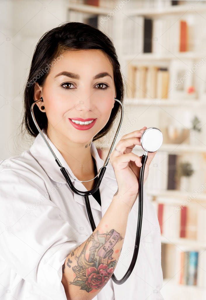Beautiful smiling tattooed young doctor holding the chest part of the stethoscope in her hand, in office background