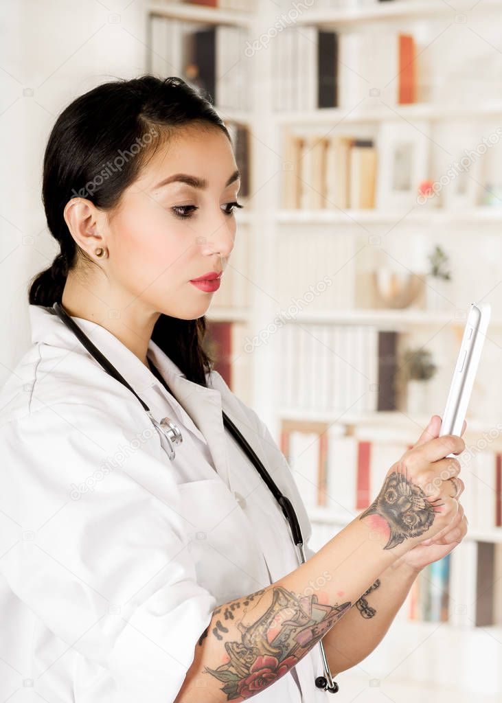Beautiful tattooed young doctor holding in her hands a tablet reading a medical results from patients, in office background