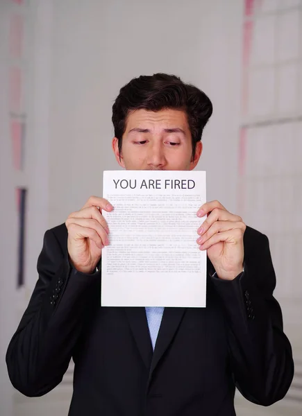 Close-up van verrast jonge zakenman dragen een pak en verschuilt zich achter een vel papier van u zijn ontslagen tekst op het, in een onscherpe achtergrond — Stockfoto