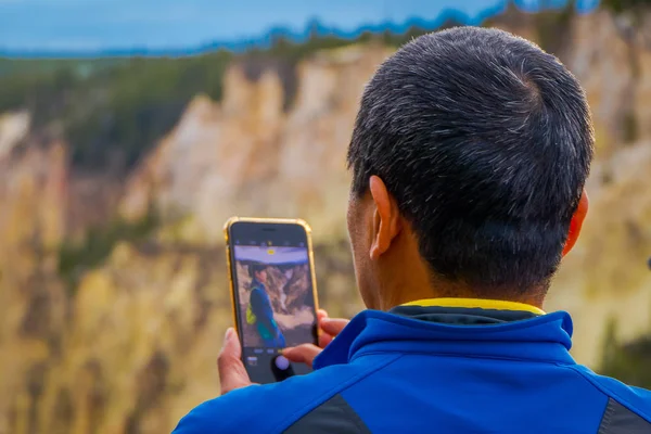 Close up of selective focus of man using a cellphone to take a picture of a friend posing in front o the upper Yellowstone Falls in Yellowstone National Park, Wyoming, United States — Stock Photo, Image