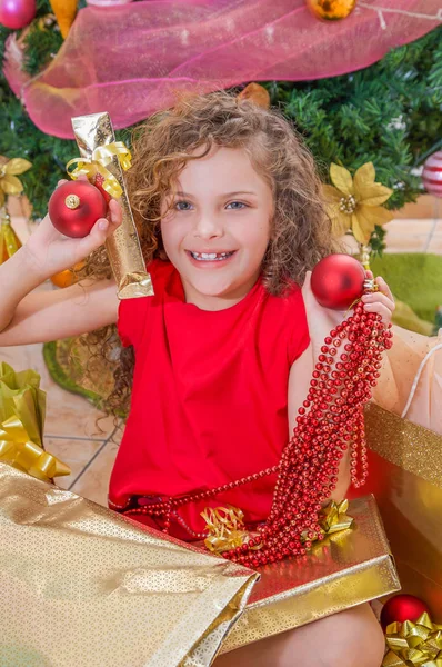 Primo piano della ragazza sorridente che indossa una camicetta rossa e tiene in mano una palla di Natale, una decorazione e un regalo, con un albero di Natale dietro, concetto di Natale — Foto Stock