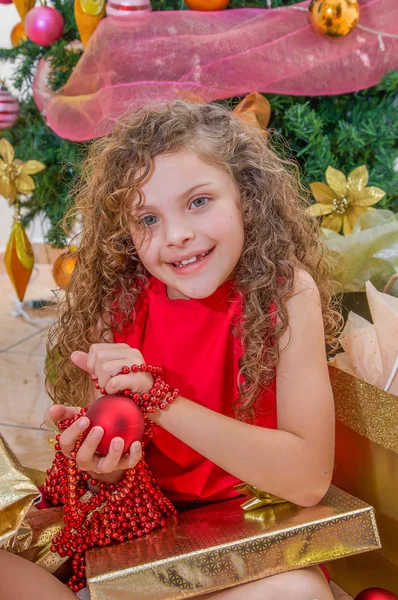 Gros plan de fille heureuse portant une robe rouge et tenant une boule de Noël et la décoration dans ses mains, avec un arbre de Noël derrière, concept de Noël — Photo