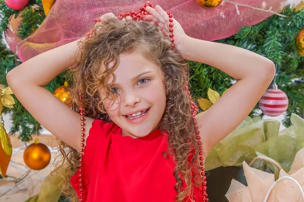 Close up van gelukkig meisje draagt een rode jurk en houdt een Kerstdecoratie in haar handen over haar hoofd, met een kerstboom achter, Kerstmis concept — Stockfoto