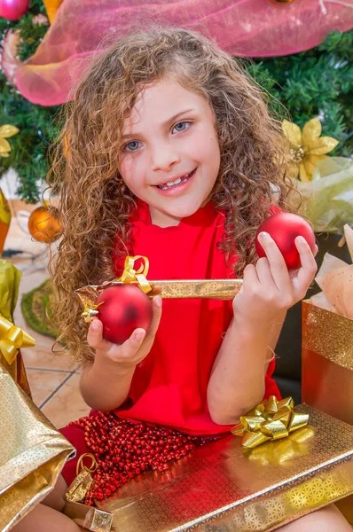 Primo piano della ragazza sorridente che indossa una camicetta rossa e tiene in mano una palla di Natale, una decorazione e un regalo, con un albero di Natale dietro, concetto di Natale — Foto Stock