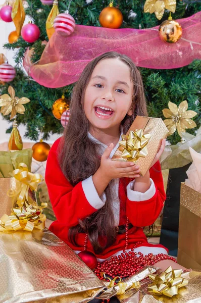 Close up van lachende meisje draagt een rode santa kostuum en bedrijf van een gift in haar handen, met een kerstboom achter, Kerstmis concept — Stockfoto