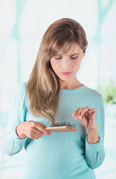 Acercamiento de una hermosa joven haciendo una cara preocupada mientras se cepilla el pelo y toma los pelos perdidos del cepillo, en un fondo borroso — Foto de Stock
