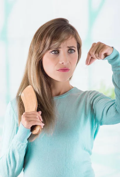 Vacker ung kvinna gör en orolig ansikte och hålla i en hand en borste medan hon håller en grupper av förlorat hår under borstning i en suddig bakgrund — Stockfoto