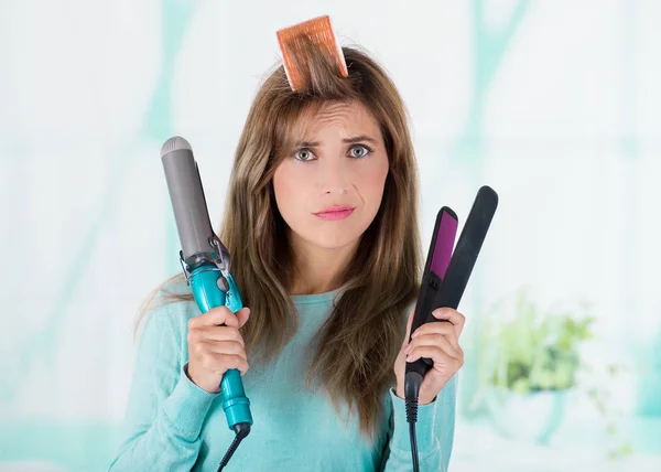 Primer plano de una mujer preocupada usando un rodillo de pelo en su cabeza y sosteniendo dos alisadores en ambas manos en un fondo borroso — Foto de Stock