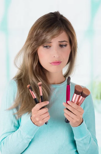 Primer plano de la joven preocupada sosteniendo en ambas manos un conjunto de pinceles de maquillaje en un fondo borroso — Foto de Stock