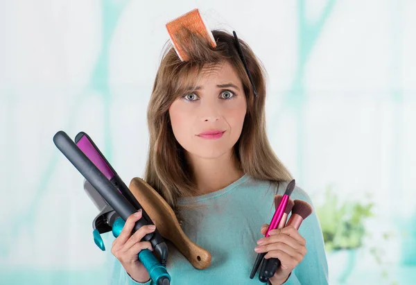 Primer plano de una joven preocupada con un rodillo de pelo en la cabeza y sosteniendo en las manos unas alisadoras y un conjunto de pinceles de maquillaje, en un fondo borroso — Foto de Stock