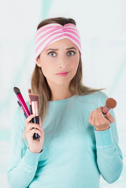 Primer plano de la joven feliz sosteniendo en ambas manos un conjunto de pinceles de maquillaje y llevando en su cabeza un pañuelo rosa y blanco, en un fondo borroso — Foto de Stock