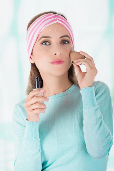 Acercamiento de una joven sexy con un pañuelo en la cabeza, sosteniendo un maquillaje y haciendo maquillaje loco en su cara usando una esponja, en un fondo borroso — Foto de Stock
