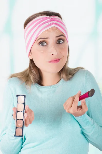 Primer plano de la joven preocupada sosteniendo una paleta de maquillaje y haciendo maquillaje loco en su cara usando un cepillo de maquillaje, en un fondo borroso — Foto de Stock